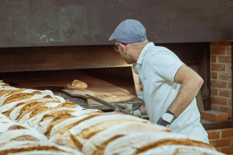 Ein Bäcker arbeitet mit dem Brotschieber am Steinofen, davor liegt eine lange Reihe frischer Brotlaibe