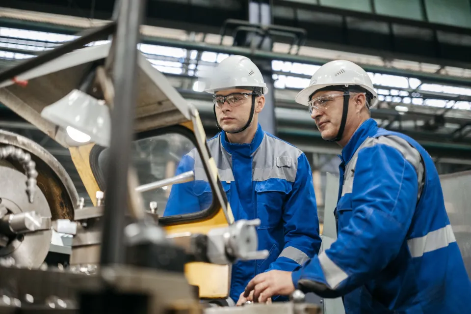 Ein erfahrener Mitarbeiter erklärt einem Kollegen an einer Maschine in der Werkshalle einen Arbeitsschritt