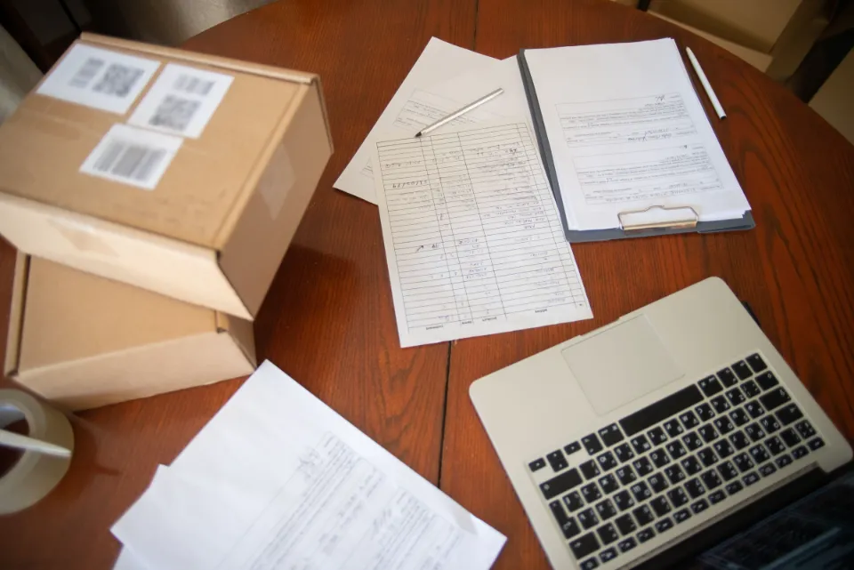Handschriftliche Formulare, ein Klemmbrett, Versandkartons und ein Laptop auf einem Holztisch