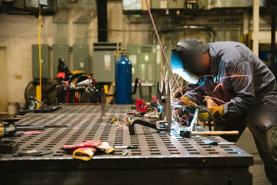 Ein Schweißer arbeitet mit Schutzhelm an einem großen Schweißtisch in einer Metallwerkstatt