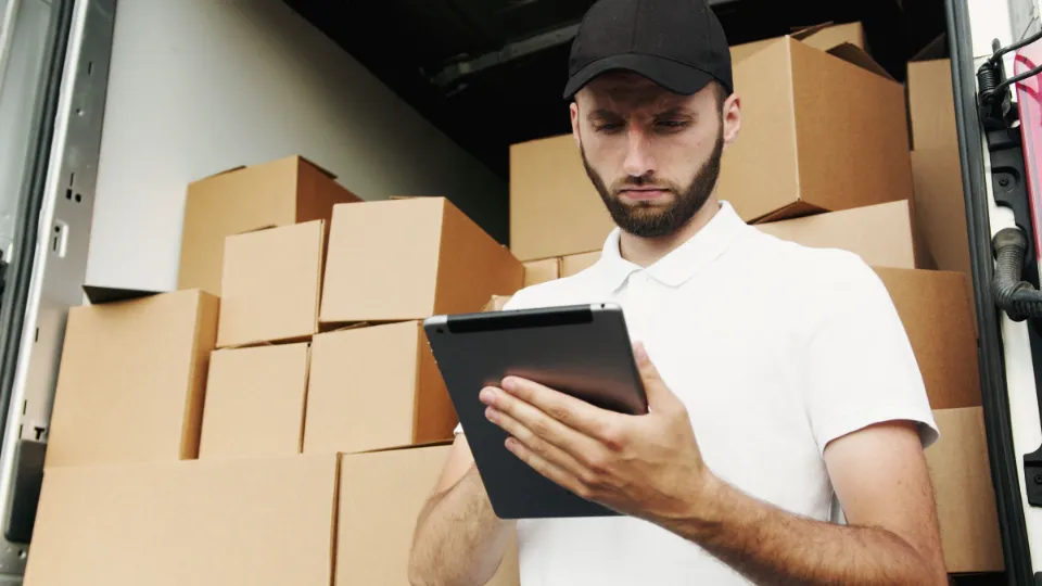 Ein Mitarbeiter prüft zwischen Kartons einen Lieferschein auf dem Tablet